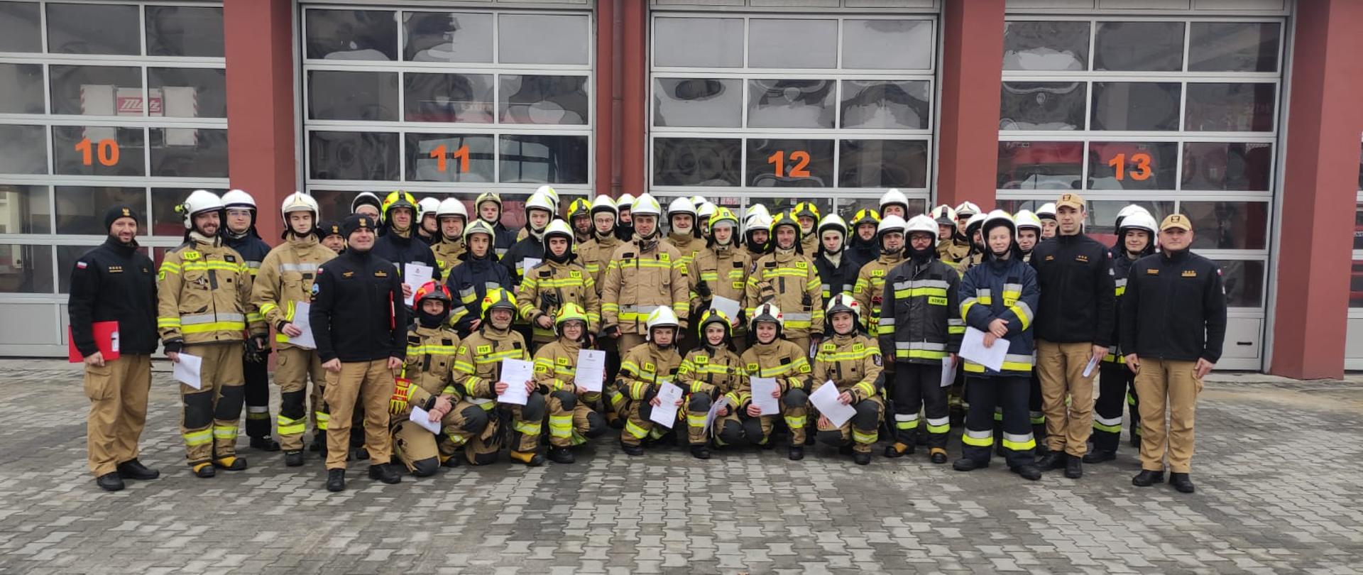 Na miejscu zbiórki strażacy OSP z ceryfikatami ukończenia szkolenia OSP. Ubrani są w mundury strażackie i znajdują się przed garażem remizy PSP.