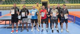 Grupa uczestników turnieju tenisa stołowego pozuje w hali sportowej z dyplomami i pucharami podczas ceremonii wręczenia nagród.