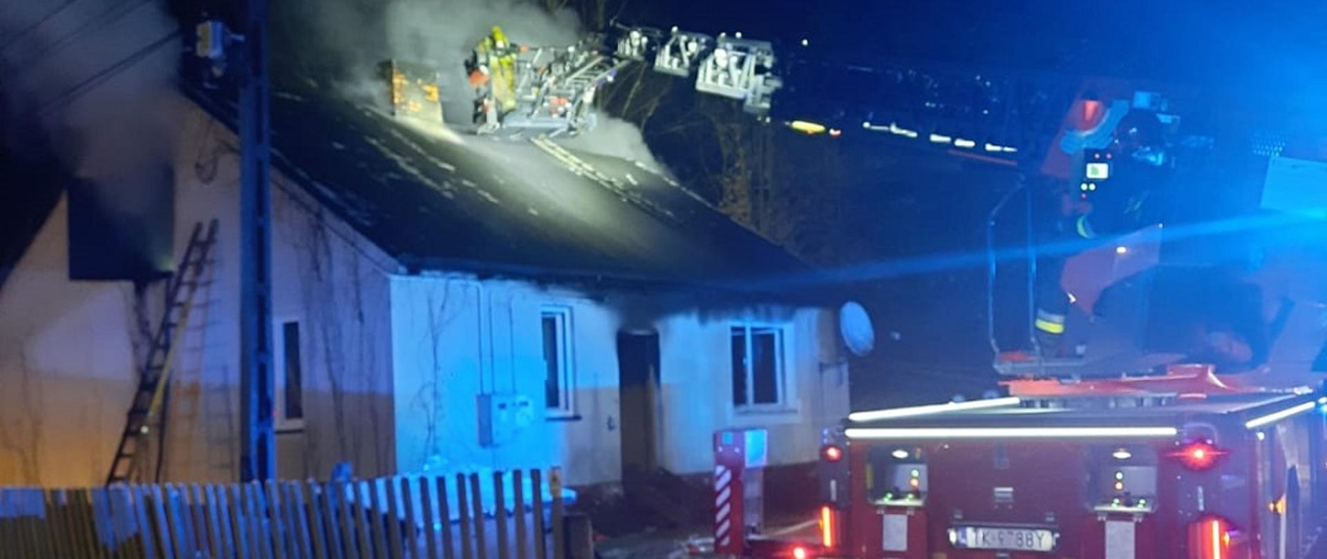 Zdjęcie obrazuje budynek murowany, parterowy. Ze strychu budynku wydobywa się dym. Przed budynkiem strażacy rozłożyli samochód z drabiną w celu wykonania otworów rewizyjnych w dachu. Pomogą one w oddymieniu strychu. 