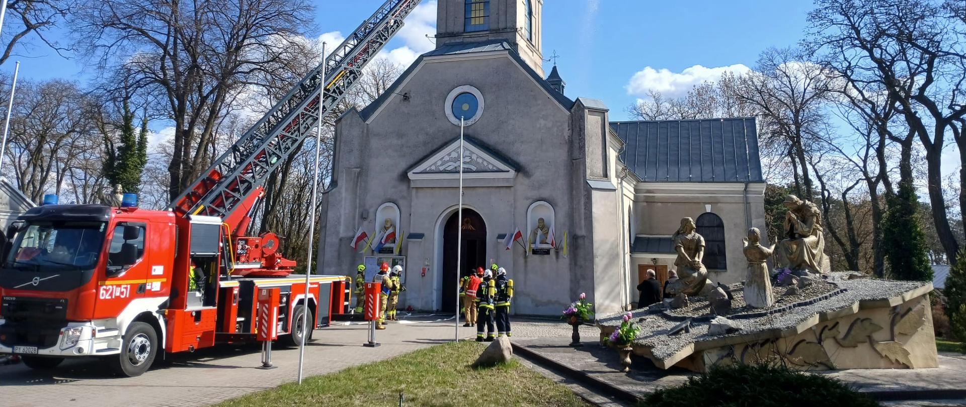 Wóz strażacki z wysuniętą drabiną stoi przed kościołem, a na jej końcu strażacy kierują strumień wody na wieżę świątyni. Przy wejściu do budynku zgromadzona jest grupa ratowników. Po prawej stronie widoczna jest rzeźba przedstawiająca scenę religijną, a wokół rosną bezlistne drzewa pod jasnym, częściowo zachmurzonym niebem.
