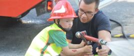 Wspieramy leczenie Gosi. 3-letni syn laureata licytacji "Dnia ze strażakami w Komendzie Powiatowej PSP w Rawiczu" w towarzystwie strażaka na placu komendy. Chłopiec ubrany w czerwony hełm i żółtą kamizelkę odblaskową wcielił się w rolę prądownika i podaje strumień wody w natarciu na "pożar".