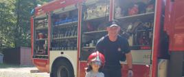 Na pierwszym planie mały chłopiec ubrany w strażacki hełm, na tle samochodu ratowniczo-gaśniczego. Chłopcu asystuje strażak.