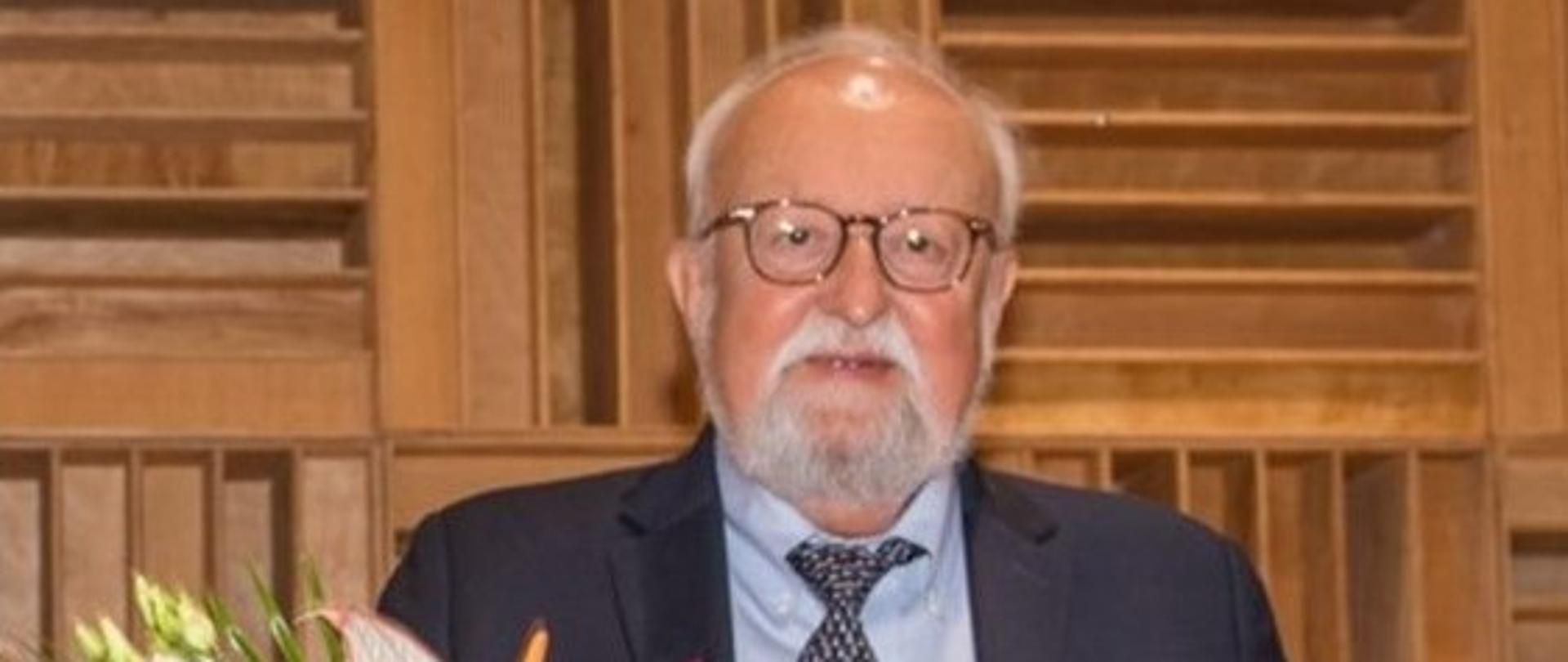 Zdjęcie przedstawiające postać kompozytora profesora Krzysztofa Pendereckiego; profesor trzyma w ręce bukiet kwiatów, które otrzymał podczas uroczystego koncertu zorganizowanego z okazji 85 urodzin K. Pendereckiego w ZPSM w Dębicy, zdjęcie zrobiono w auli szkoły. 