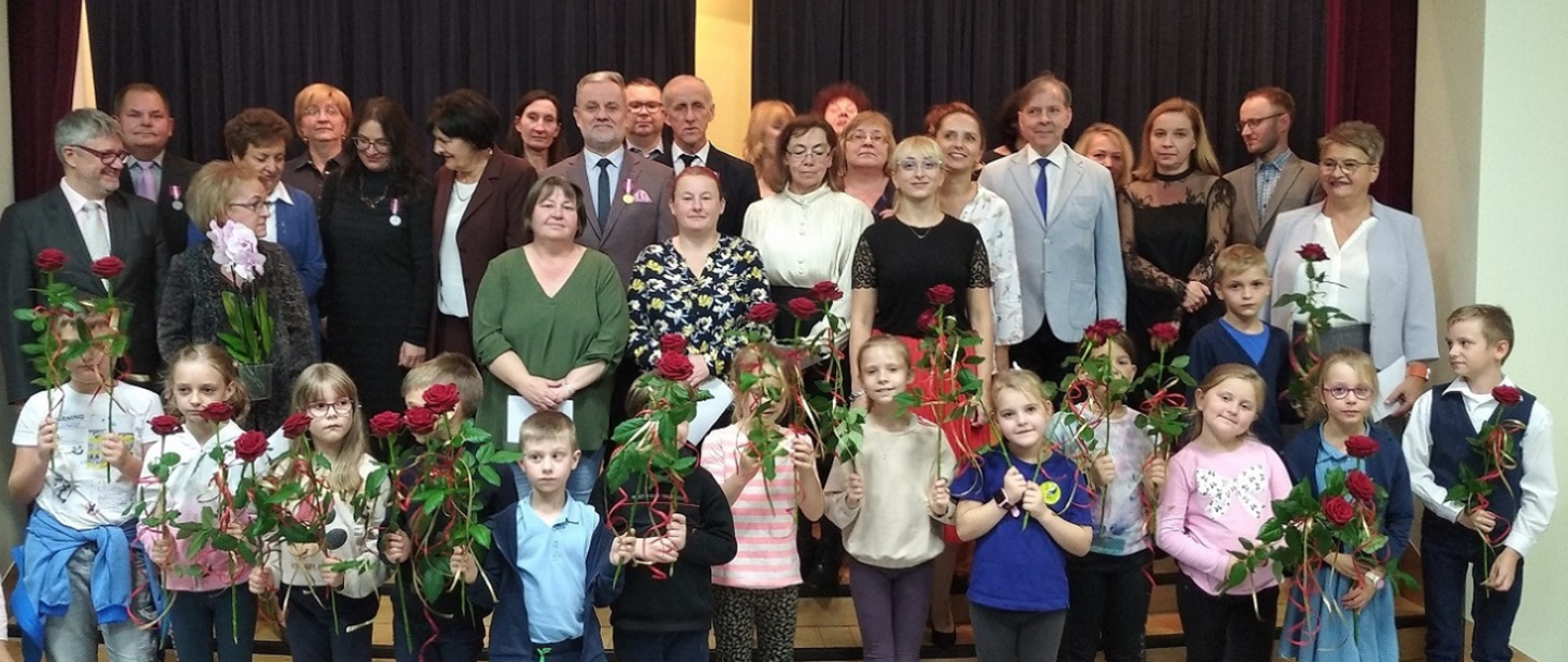 Zdjęcie przedstawia osoby ustawione w trzech rzędach na scenie w sali koncertowej szkoły. W pierwszym rzędzie stoją małe dzieci z różami czerwonymi w rękach. W drugim i trzecim rzędzie, na schodach sceny, stoją nauczyciele i pracownicy szkoły odświętnie ubrani w radosnych nastrojach.