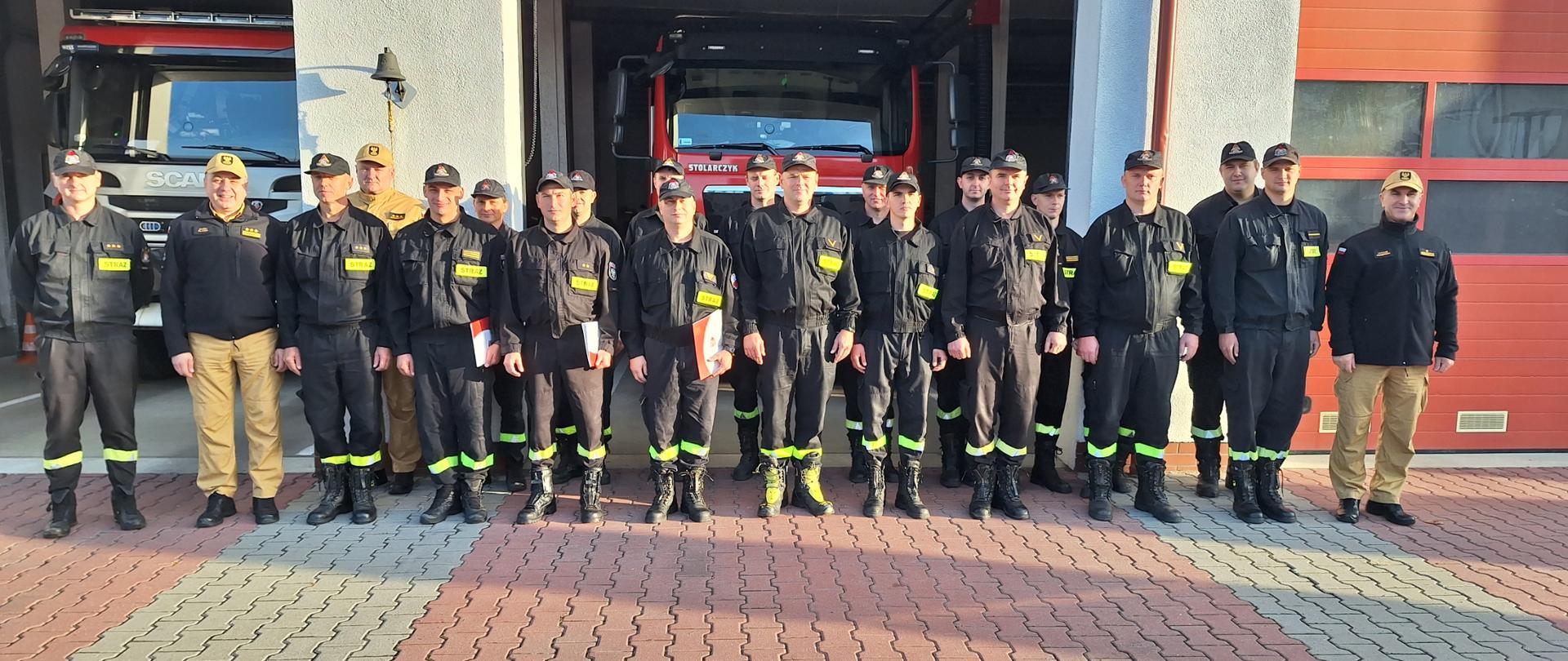 Zdjęcie przedstawia strażaków stojących w dwuszeregu przed budynkiem strażnicy. Bramy garażu podniesione do góry. Z tyłu widać samochody strażackie