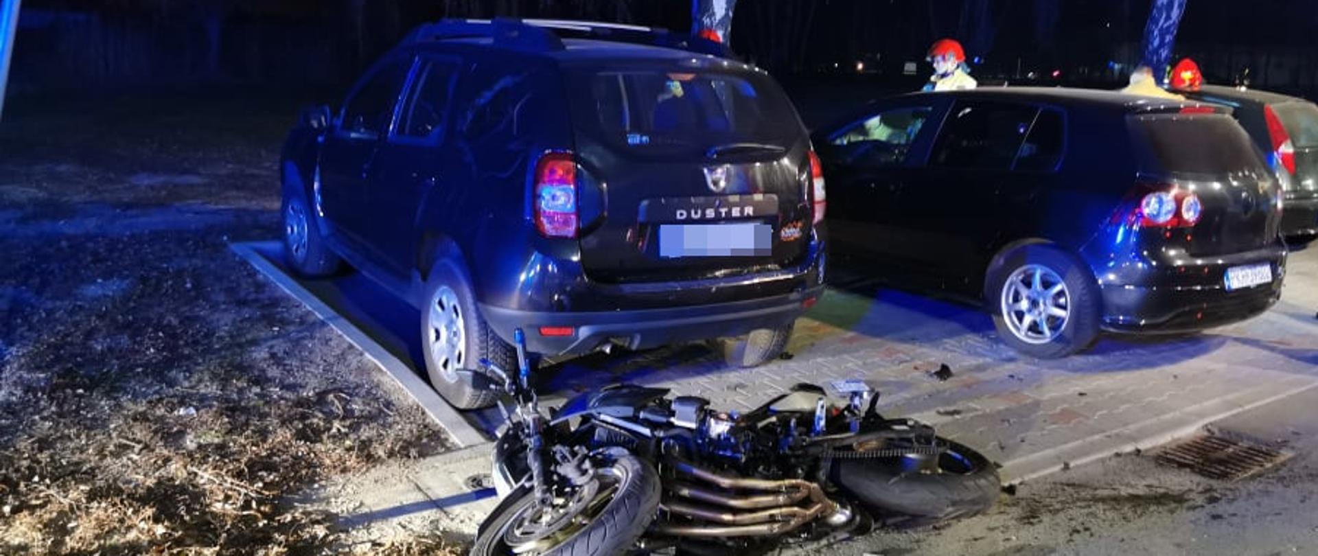 Przy parkingu, na którym stoją zaparkowane samochody osobowe leży uszkodzony motocykl . W tle widać strażaków realizujących działania.
