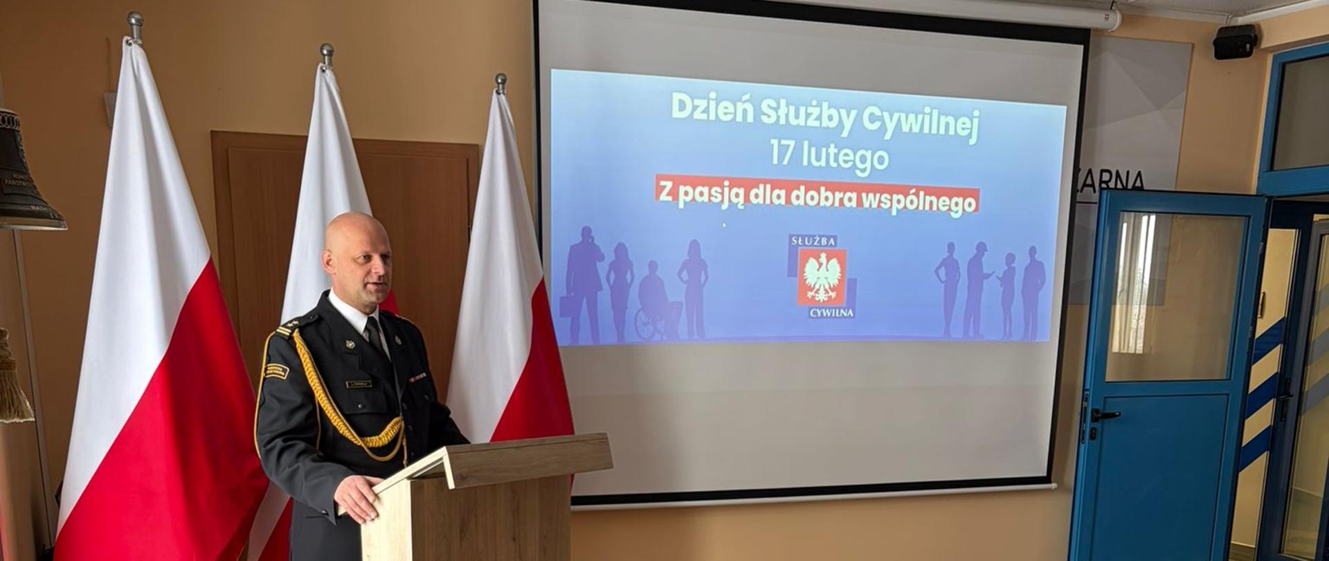 Komendant Powiatowy Państwowej Straży Pożarnej w Lęborku przemawia przy drewnianej mównicy z logo Państwowej Straży Pożarnej. Na stoliku przykrytym zielonym materiałem, znajdującym się z przodu mównicy na czerwonej tacy leżą okolicznościowe medale i odznaczenia oraz kosze prezentowe i torba papierowa. Na ekranie do projektora za komendantem wyświetlony na niebieskim tle napis Dzień Służby Cywilnej 17 lutego Z pasją dla dobra wspólnego oraz logo służby cywilnej.
