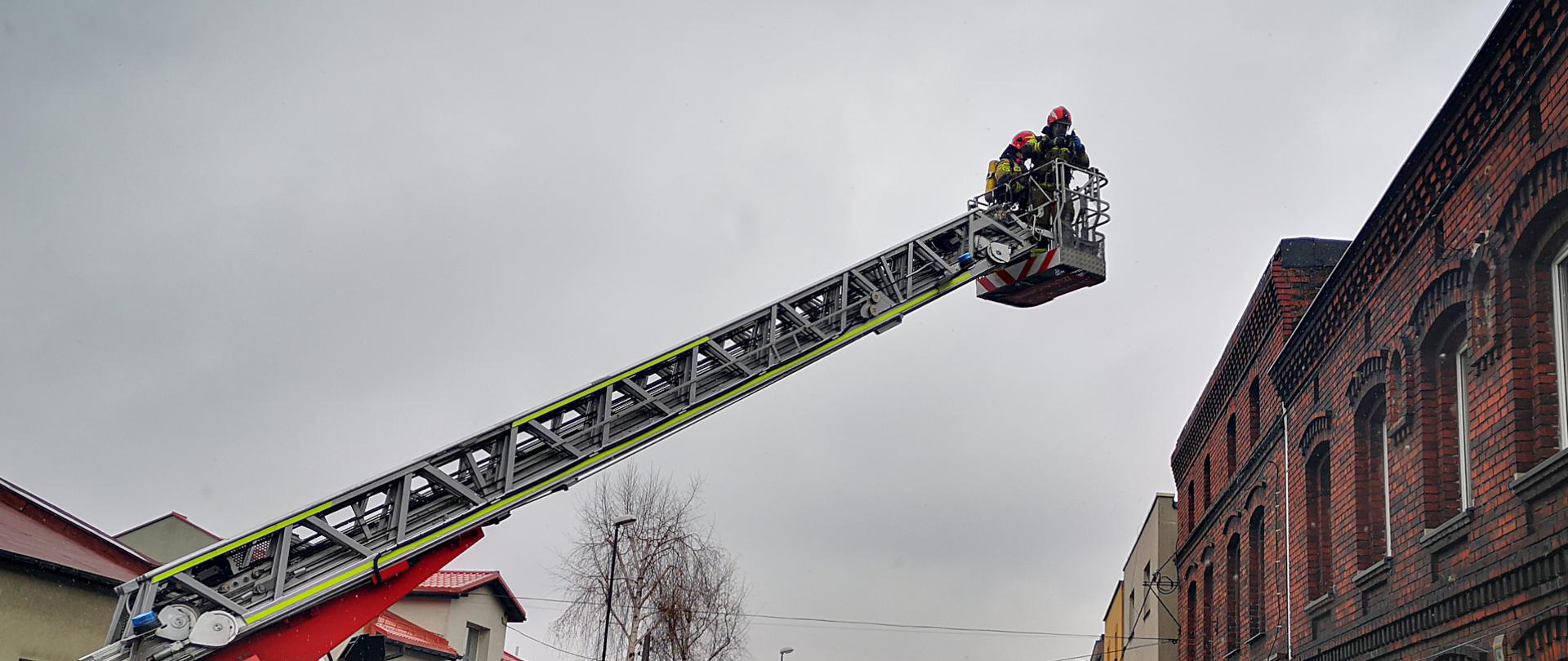Zdjęcie przedstawia dwóch ratowników w ubraniach specjalnych znajdujących się w koszu drabiny mechanicznej, zbliżających się się do dachu budynku wielorodzinnego.