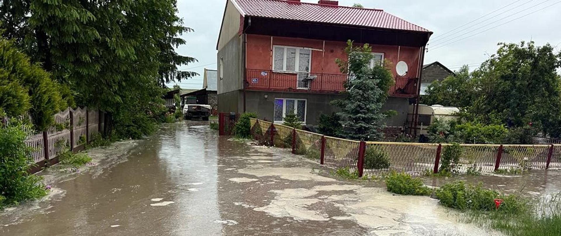 Na zdjęciu budynek mieszkalny piętrowy. Teren posesji, na której znajduję się dom częściowo zalany jest wodą. 