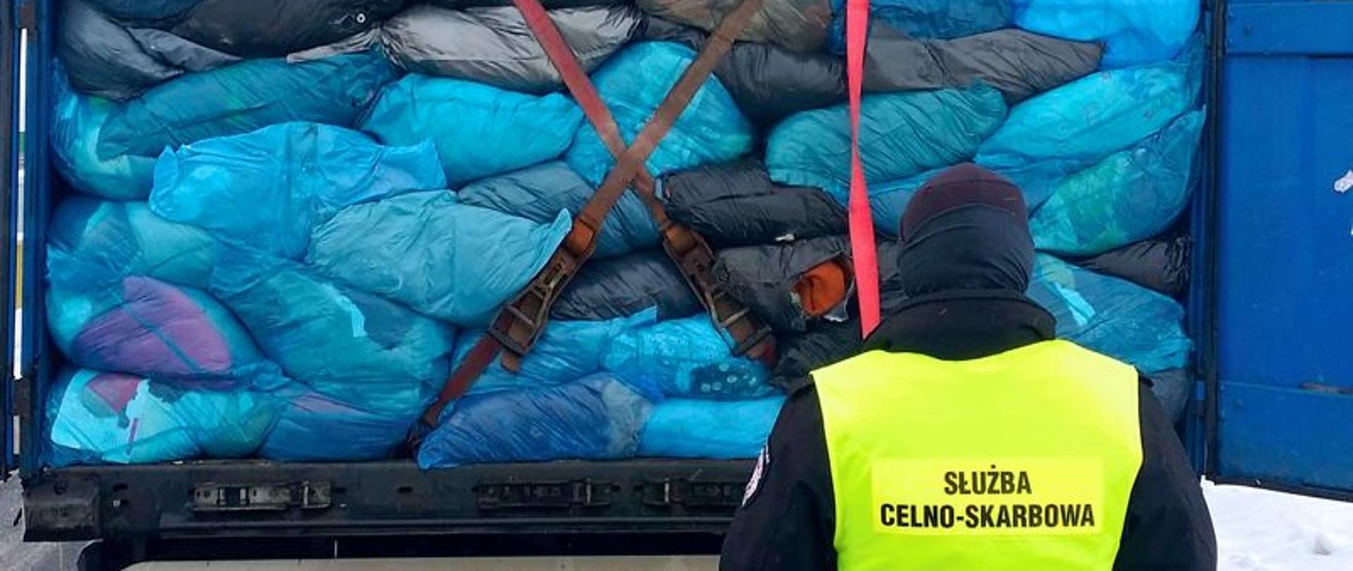Funkcjonariusz Służby Celno-Skarbowej z tyłu otwartej naczepy samochodu ciężarowego, naczepa wypełniona niebieskimi workami.