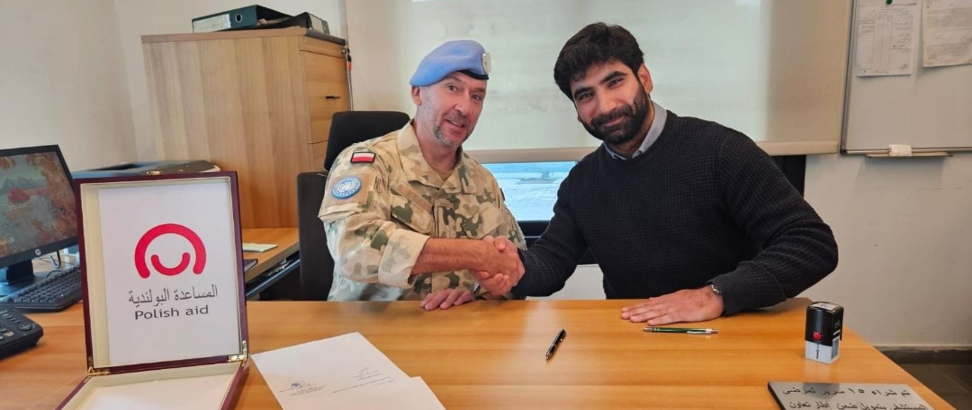 Dwóch mężczyzn ściska sobie rękę przy biurku pozując do zdjęcia, jeden ma błękitny beret i mundur w polskim kamuflażu z polską flagą, przed nimi pudełko na dokumenty z logo Polska pomoc i podpisana umowa