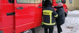 Strażak pomaga osobie starszej wsiąść do samochodu strażackiego podtrzymując ją za rękę. Kobieta drugą ręką pomaga sobie chwytając pojazdu.