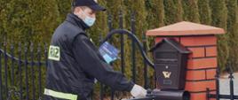 Strażak w umundurowaniu koszarowym stoi przed ogrodzeniem posecji i wkłada prawą ręką ulotkę do skrzynki listowej. Na rękach ma rękawiczki jednorazowe i na twarzy maseczkę. w drukiej ręce trzyma plik ulotek. W tle szpaler tui.