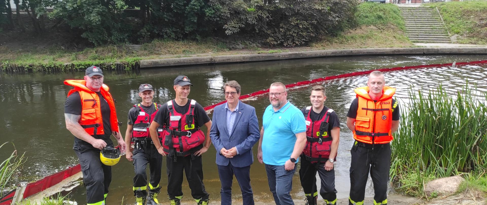 Na tle rzeki, na której jest rozstawiona czerwona zapora stoją ustawieni strażacy w kapokach ratunkowych oraz przedstawiciele samorządu, starosta braniewski i pracownik obony cywilnej. W oddali drugi brzeg rzeki i zielone drzewa.