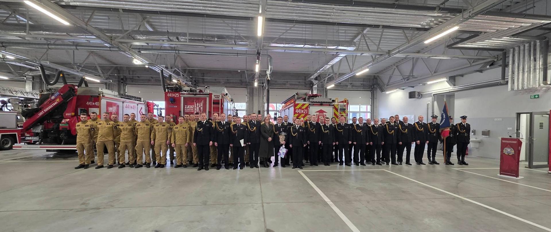 Pamiątkowe zdjęcie strażaków w piaskowych i galowych mundurach w hali garażowej. W tle wozy strażackie.
