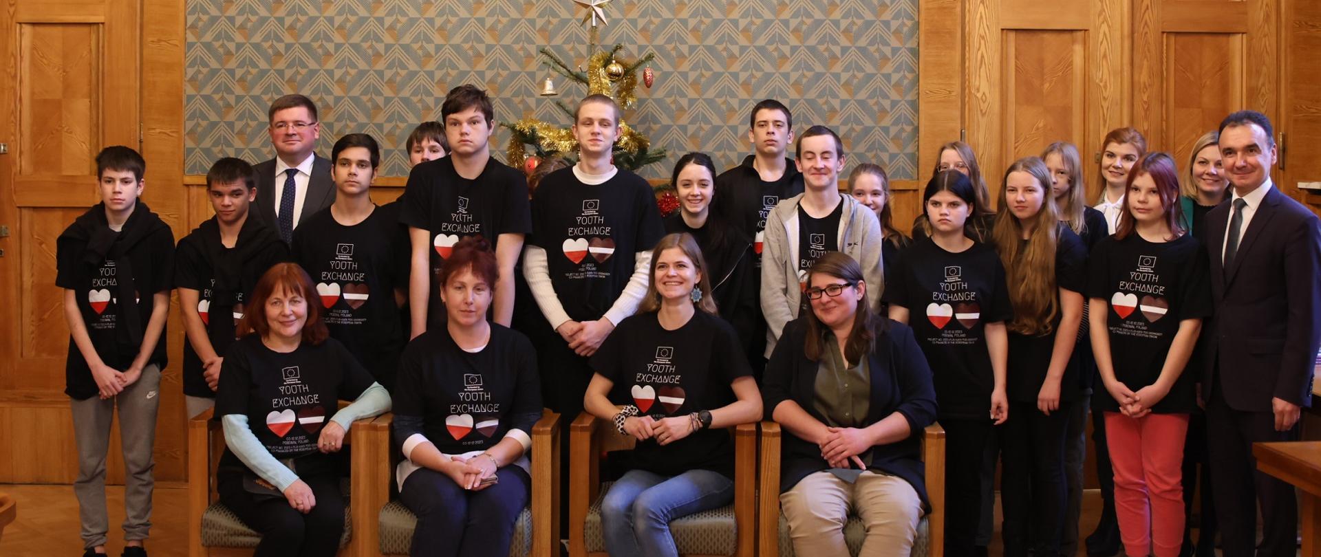 W sali ze ścianami wyłożonymi drewnianymi panelami stoi duża grupa młodzieży w czarnych koszulkach, kilkoro z przodu siedzi na drewnianych krzesłach.