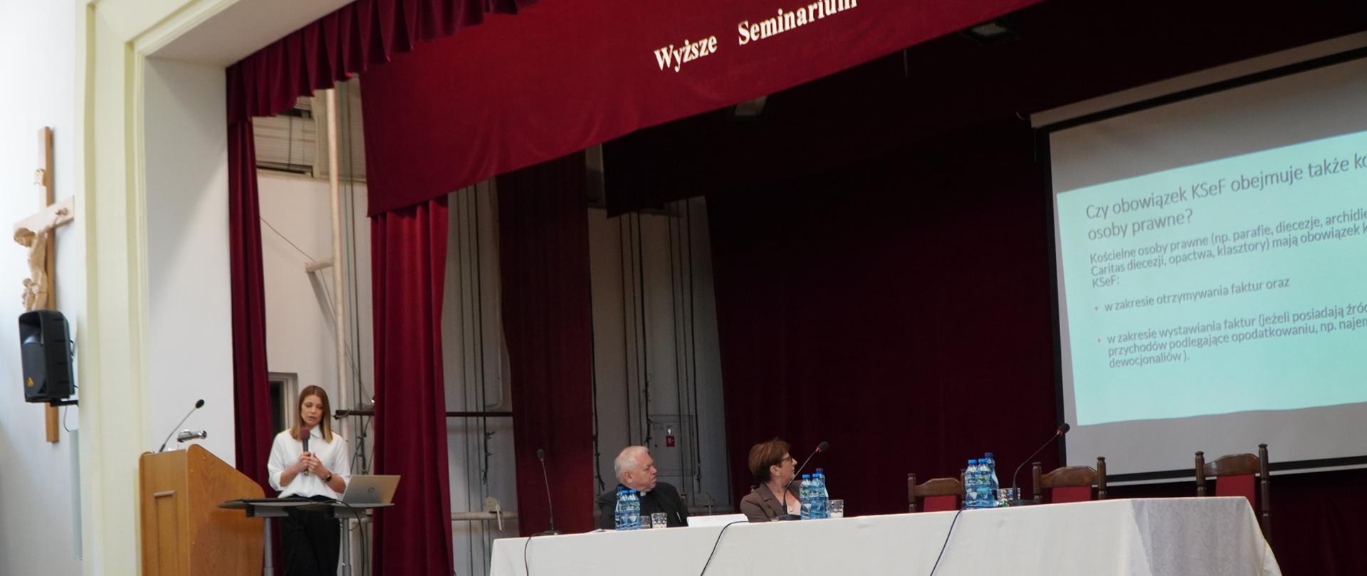 Przy mównicy stoi kobieta z mikrofonem w ręce. Obok przy stole siedzi druga kobieta i mężczyzna