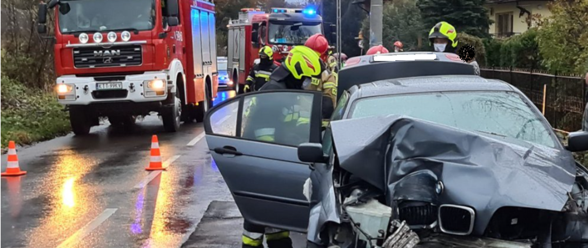 Zdjęcie przedstawia wypadek samochodu, który wypadł z zakrętu i uderzył w słup energetyczny. W skutek uderzenia samochód został mocno zniszczony. W tle widać strażaków i wozy strażackie zabezpieczające miejsce zdarzenia.