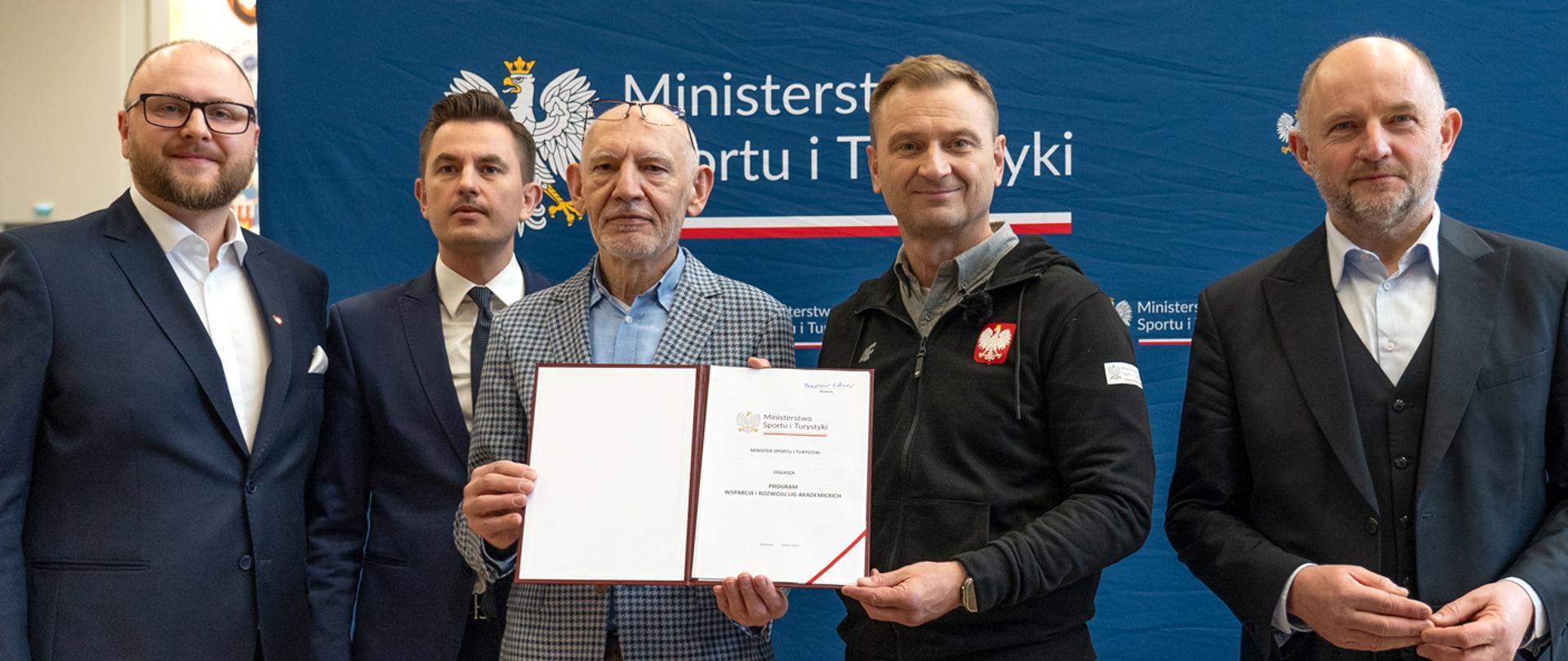Minister Sportu i Turystyki ogłasza Program Wsparcia i Rozwoju Lig Akademickich - na zdjęciu minister Sławomir Nitras wraz z uczestniczącymi w konferencji mężczyznami prezentuje podpisany dokument; w tle ścianka z logotypami ministerstwa