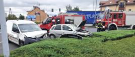 Widać dwa uszkodzone samochody, które są na poboczu, za nimi dwa samochody straży i kilku strażaków