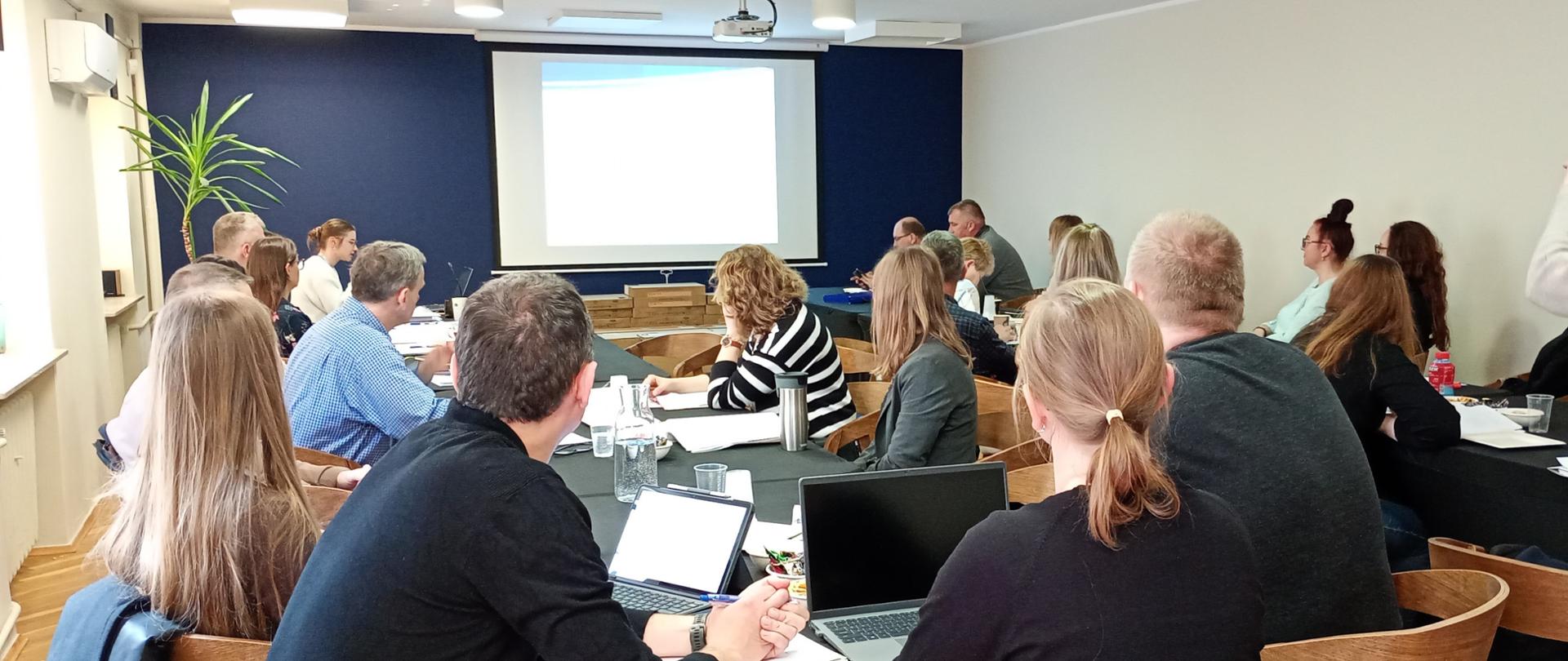 Uczestnicy szkolenia siedzą w sali konferencyjnej patrzą na ekran w głębi sali