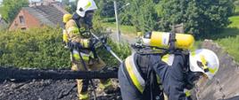 Dwóch strażaków w aparatach powietrznych pracuje na spalonym dachu budynku. Jeden z nich trzyma narzędzie do rozbiórki, drugi operuje wężem gaśniczym.