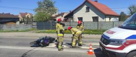 Czterech strażaków usuwa plamę oleju z jezdni obok leży przewrócony motocykl.