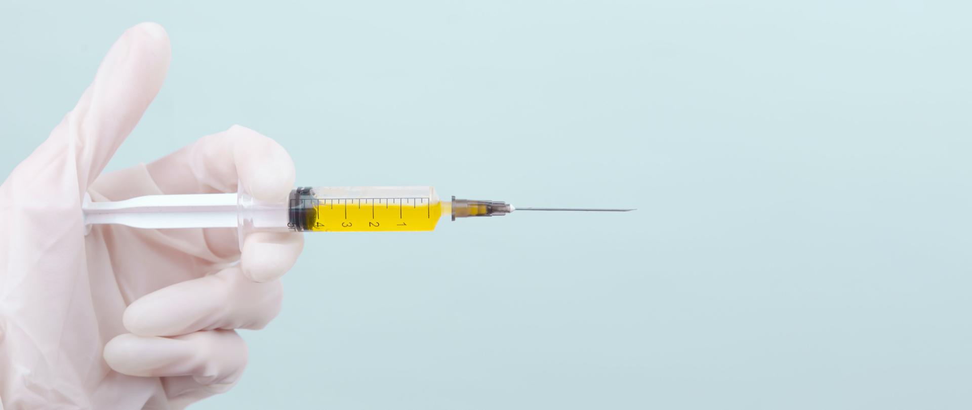 Zdjęcie przedstawia trzymaną w dłoni strzykawkę wraz ze szczepionką. 