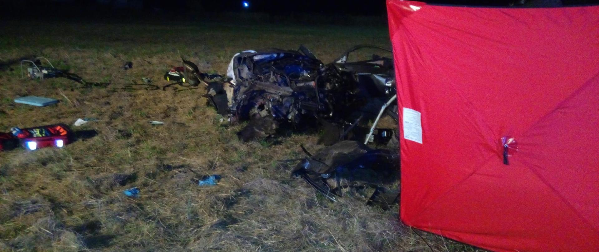 Zdjęcie przedstawia miejsce wypadku: z prawej strony rozstawiono czerwony parawan zza którego wystają fragmenty zniszczonego auta.