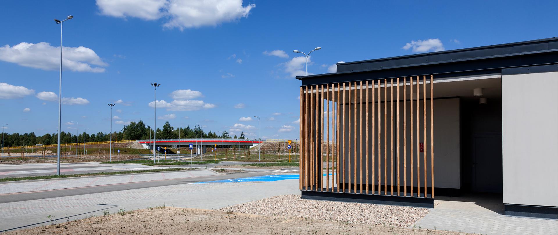 Fotografia Miejsca Obsługi Podróżnych Woźniki Wschód na A1 w woj. śląskim. W lewej części fotografii widoczny budynek sanitarnym. Środkową cześć fotografii zajmują drogi manewrowe i miejsca parkingowe wyposażone w oznakowanie oraz oświetlenie drogowe. W centralne części fotografii widoczny obiekt drogowy nad autostradą łączący parę MOP-ów. 