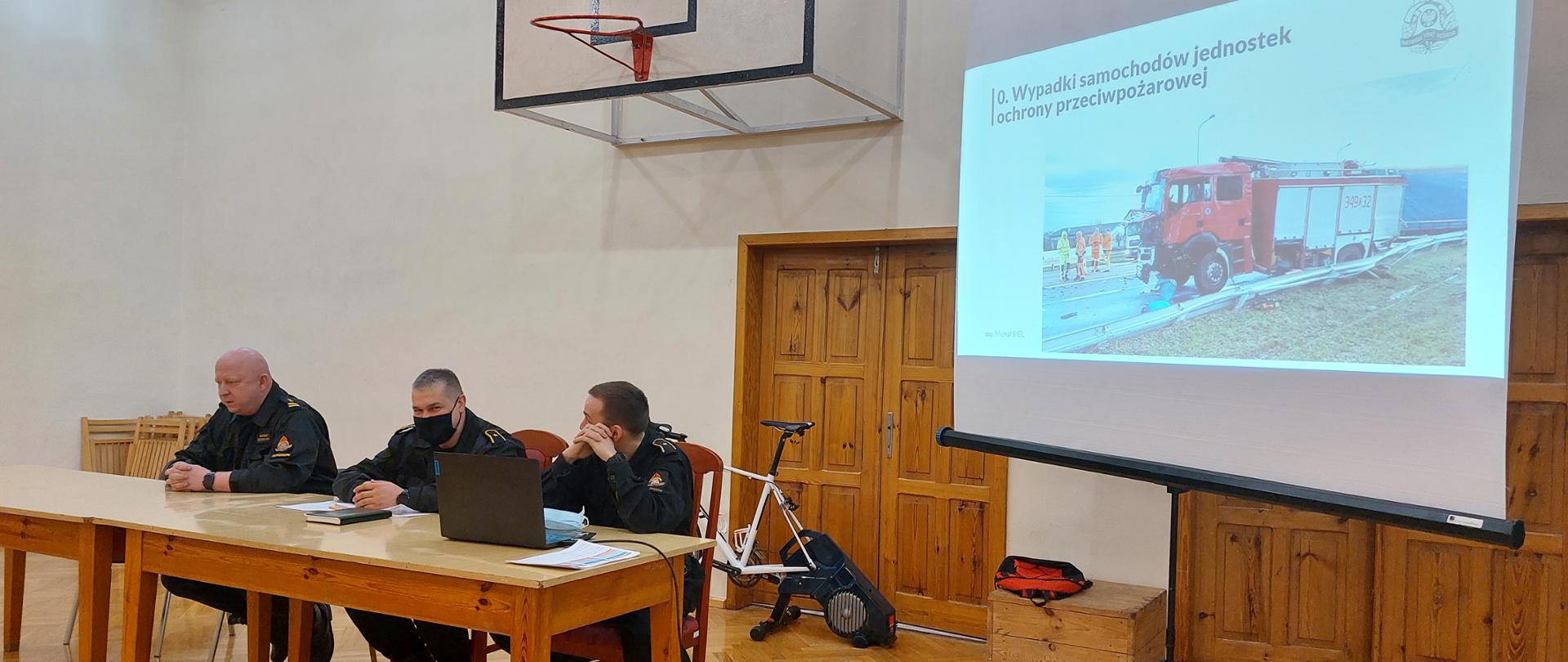 Zdjęcie wykonane w trakcie spotkania z naczelnikami. Po lewej stronie siedzą przy stole prelegenci. Po prawej stronie ekran projektora, na którym wyświetlana jest tematyczna prezentacja.