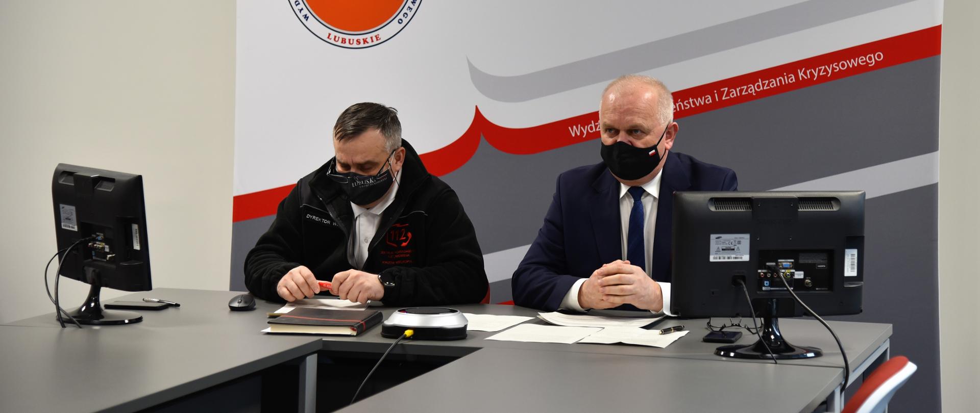 Wojewoda z dyrektorem siedzą na tle ścianki promocyjnej wydziału bezpieczeństwa i zarządzania kryzysowego. Na rogach stołu stoją dwa monitory, na środku zestaw konferencyjny. Na stole leżą również dokumenty i notatki