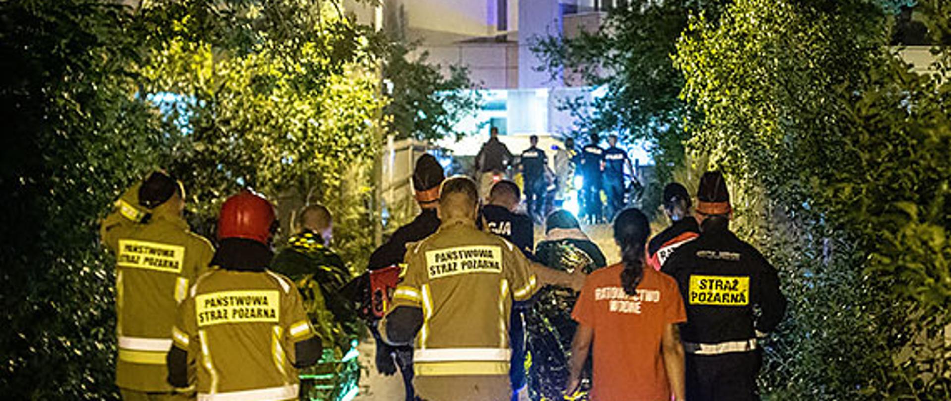 Zdjęcie przedstawia służby ratownicze prowadzące poszkodowanych chodnikiem.w tle widać budynek.