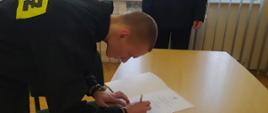 W sali konferencyjnej strażak w czarnym mundurze koszarowym składa podpis na dokumencie, komendy komendant powiatowy w tle ubrany w czarny mundur wyjściowy.
