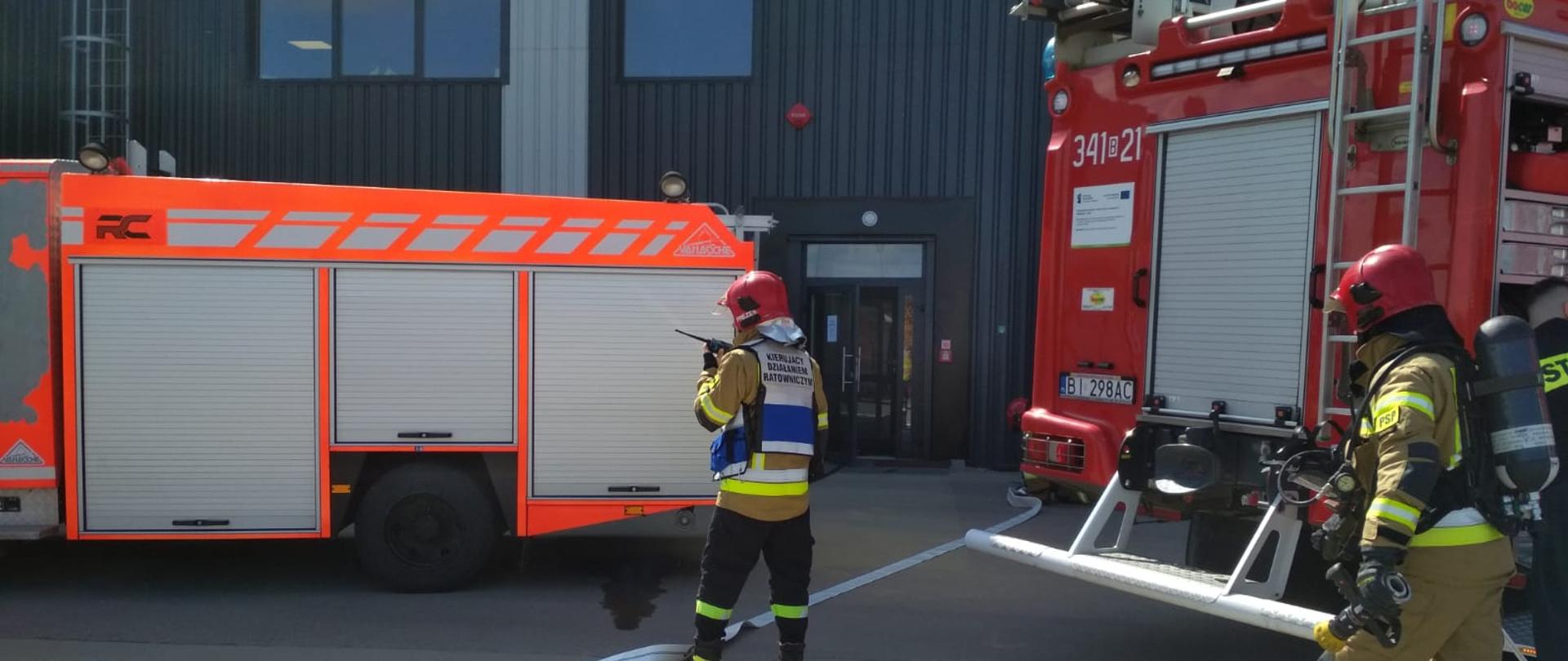 Zdjęcie przedstawia KDR wraz samochodami pożarniczymi przed wejściem do budynku hali produkcyjnej