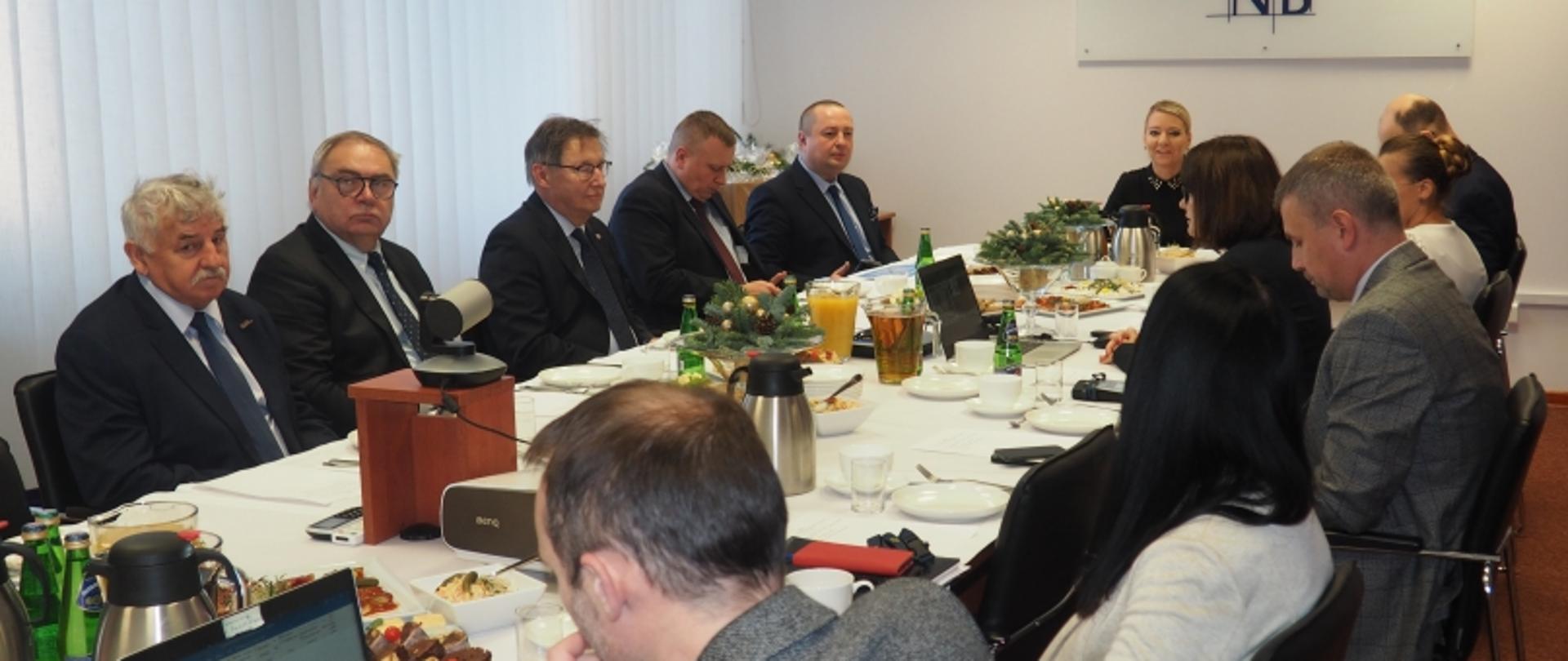 Spotkanie przy stole konferencyjnym z jedzeniem, laptopami i świątecznymi ozdobami, uczestnicy w trakcie rozmowy.