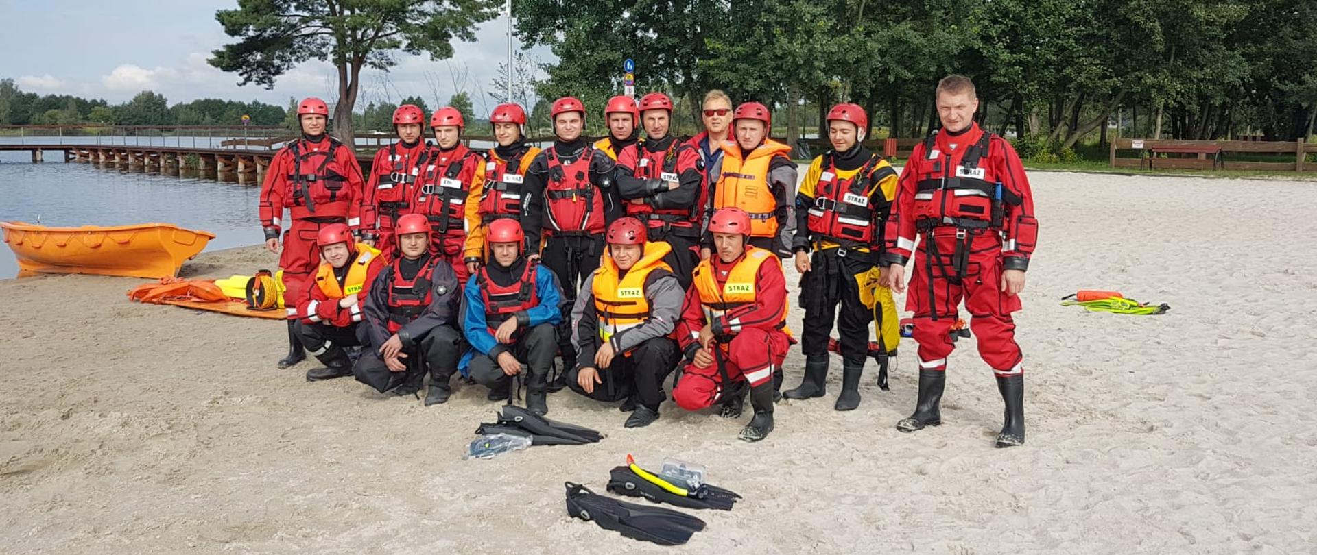 Wspólne zdjęcie grupowe strażaków w ubraniach wypornościowych na plaży jednego z kąpielisk. Strażacy przygotowani do ćwiczeń w ramach zajęć z ratownictwa wodnego.