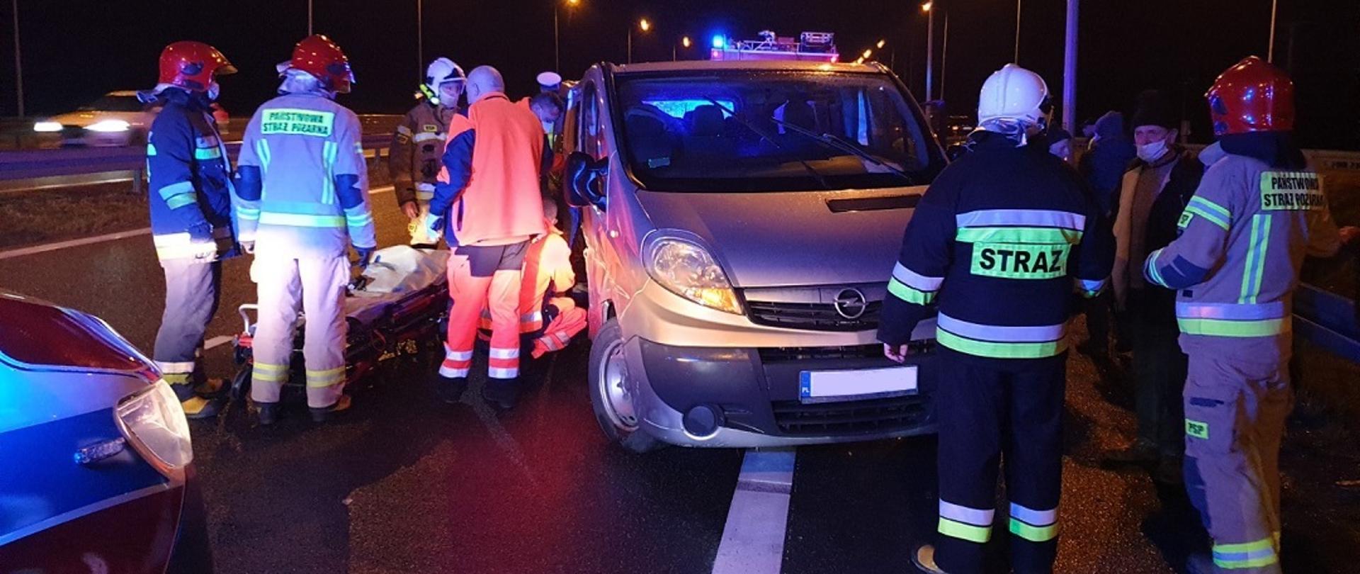 Zdjęcie obrazuje samochód marki Opel Vivaro, który stoi na pasie rogi ekspresowej S-7. Uderzył on wcześniej w amortyzator energochłonny znajdujący się przy zjeździe na Suchedniów. Wokół pojazdu znajdują się ratownicy PSP. Z lewej przy samochodzie znajduje się dwóch ratowników medycznych, którzy udzielają pomocy poszkodowanemu. W tle zdjęcia widzimy nosze, gotowe do zabrania poszkodowanego do karetki.