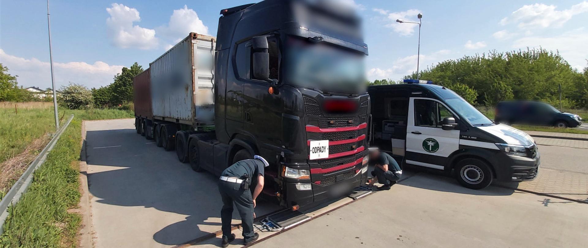 Ciężarówka stojąca przez wagą w nawierzchni zatoki. Obok stoją dwaj inspektorzy.