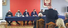 Zdjęcie wykonane w Sali szkoleniowej ochotniczej straży pożarnej w Goździe. Na zdjęciu widać na środku sali ustawiony stół przy którym siedzą: komendant miejski Państwowej Straży Pożarnej w Radomiu brygadier Paweł Fliszkiewicz, wójt gminy Gózd Paweł Dziewit, starosta radomski Waldemar Trelka, Wicewojewoda mazowiecki Artur Standowicz, Zastępca Mazowieckiego Komendanta Wojewódzkiego Państwowej Straży Pożarnej starszy brygadier Artur Gonera oraz zastępca komendanta miejskiego Państwowej Straży Pożarnej w Radomiu brygadier Bartosz Musiał. Frontem do osób siedzących przy stole ustawione są krzesła na których siedzą przybyli zainteresowani. Jedna z osób stoi i zadaje pytanie. Zdjęcie wykonane z bliższej perspektywy do poprzedzającego.