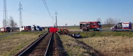 Przejazd kolejowy, samochód osobowy leży u podnóża nasypu kolejowego. Na jezdni ustawione samochody służ uczestniczących w działaniach ratowniczych. Przy samochodzie na torach kolejowych strażacy stojący obok parawanu. 