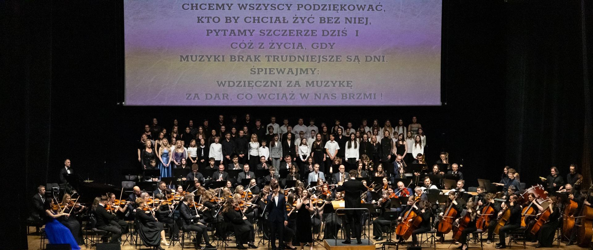 Na scenie wykonawcy - ponad 150 chórzystów i instrumentalistów. Na przodzie soliści. Nad sceną na ekranie tekst refrenu piosenki