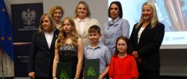 Troje dzieci pozuje do zdjęcia. Za dziećmi stoją przedstawiciele instytucji organizujących konkurs na rymowankę. Dzieci są laureatami konkursu. To Róża Pawłowska, Mikołaj Piotrowski i Amelia Marcinkowska. Wszystkie osoby się uśmiechają.