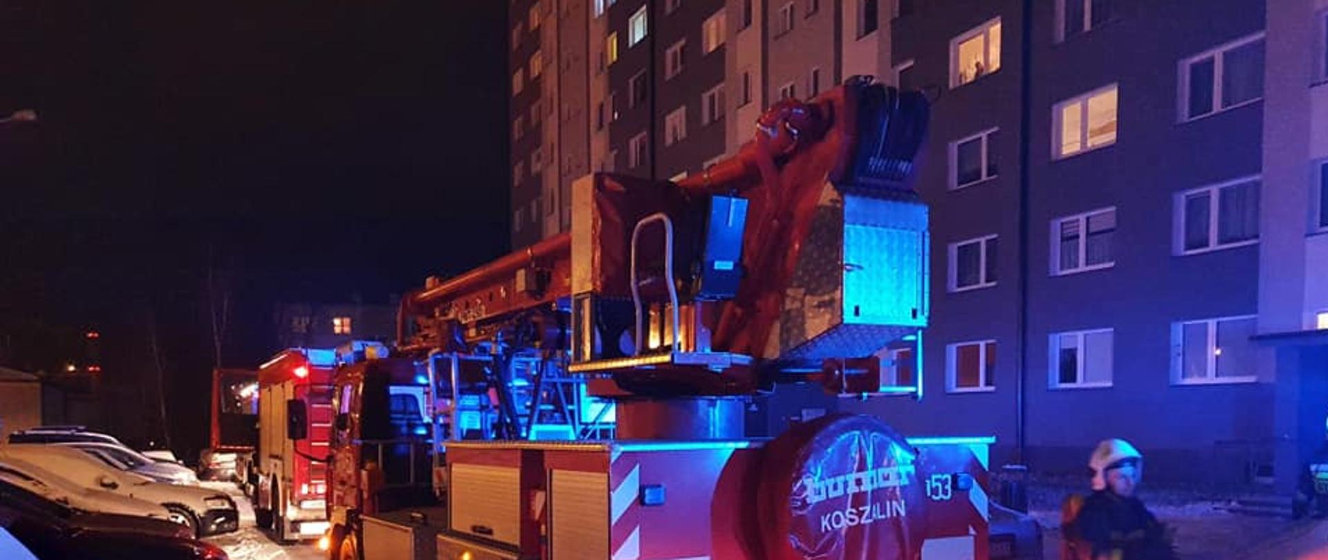 Na zdjęciu przedstawiony jest parking przed blokiem, w którym doszło do pożaru. Na parking wieżdżają dwa pojazdy straży pożarnej. Jeden gaśniczy drugi drabina. Z prawej strony widać część bloku wielorodzinnego. Na parkingu widoczne są zaparkowane pojazdy osobowe.