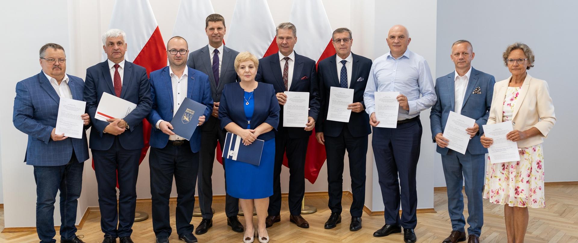 Zdjęcie zbiorowe, grupa ludzi stoi pod ścianą, pokazują trzymane w rękach dokumenty.