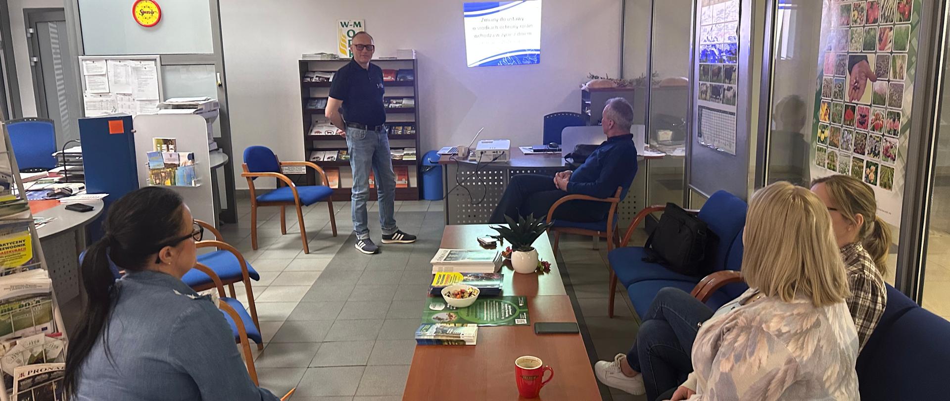 Grupa osób siedzi przy stolikach po prawej stronie. W tle na ścianie wyświetlany tytuł prezentacji : zmiany w przepisach ustawy o środkach ochrony roślin.