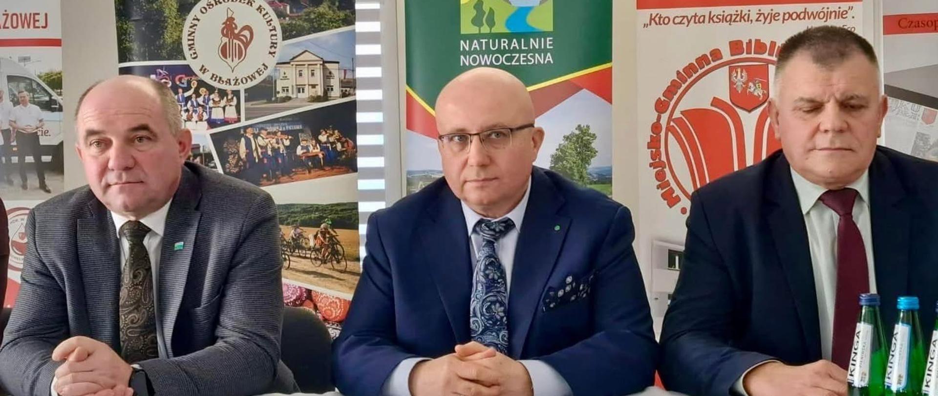Trzech mężczyzn w garniturach siedzących przy stole. Stół przykryty jest białym obrusem. Za plecami mężczyzn znajdują się banery Gminnego Ośrodka Kultury w Błażowej, Gminy Błażowa oraz Miejsko-Gminnej Biblioteki Publicznej w Błażowej.