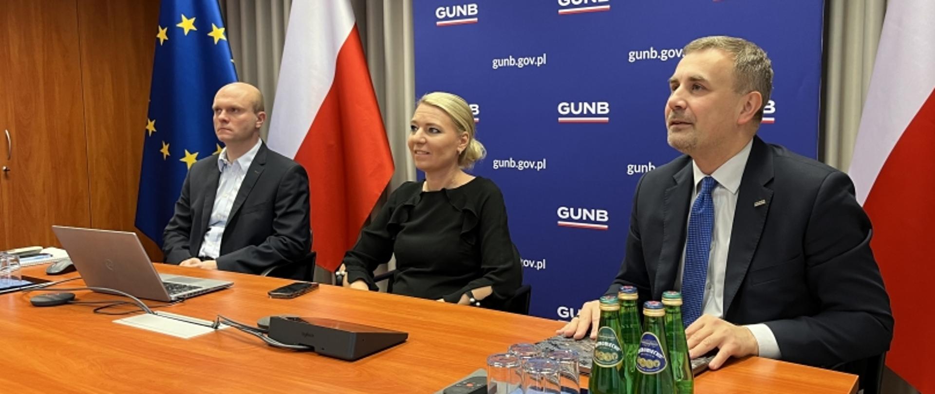 Trzy osoby przy stole konferencyjnym, za nimi baner GUNB oraz flagi UE i Polski.