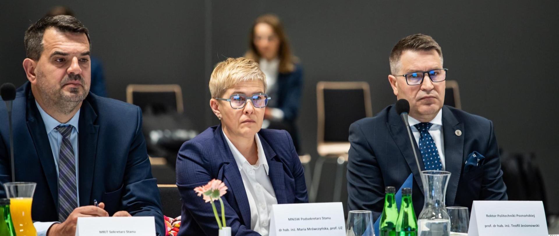 Wiceminister Maria Mrówczyńska siedzi za stołem, obok niej dwóch mężczyzn w garniturach.