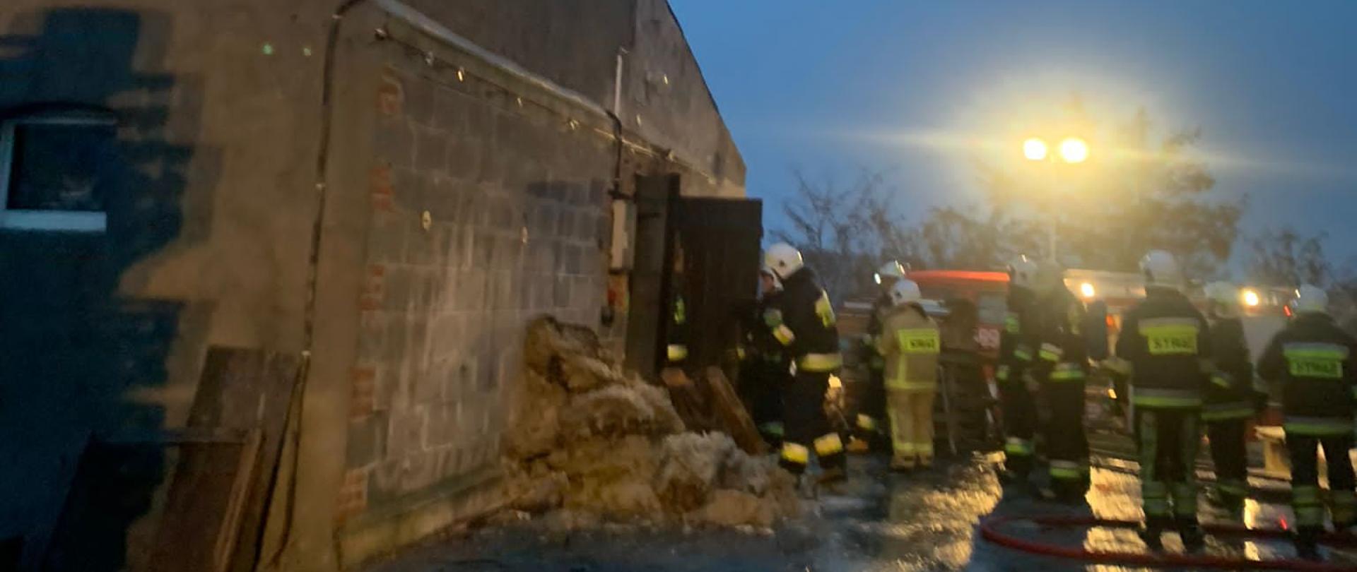 Budynek przy którym pracują strażacy przy ścianie leży niewielka ilość słomy przed budynkiem rozwinięta linia wężowa